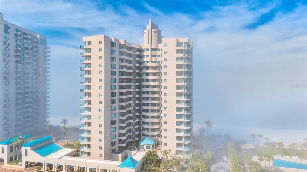An aerial view shows a tall, multi-story residential building surrounded by fog and palm trees — 1520 GULF BOULEVARD UNIT 1807, CLEARWATER BEACH, FL
