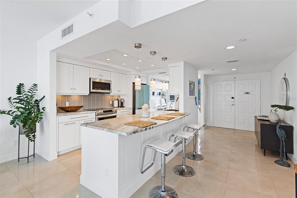 A modern kitchen features white cabinetry, a marble-topped island with bar stools, and tile flooring — 1520 GULF BOULEVARD UNIT 1807, CLEARWATER BEACH, FL