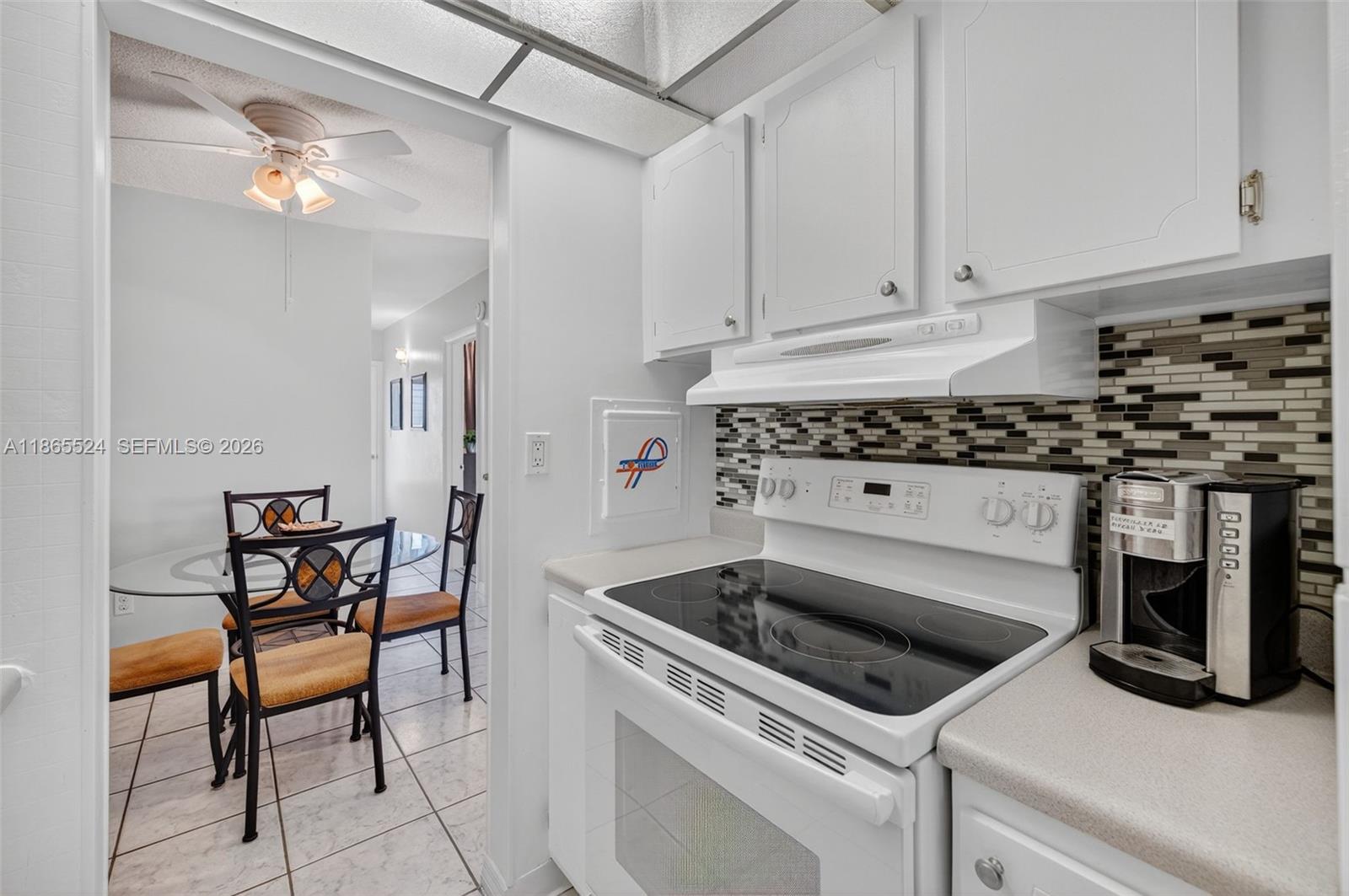 A white kitchen with a glass-top stove and mosaic tile backsplash overlooks a dining area — 16901 NE 13th Ave # 210, Miami FL 33162, MIAMI, FL