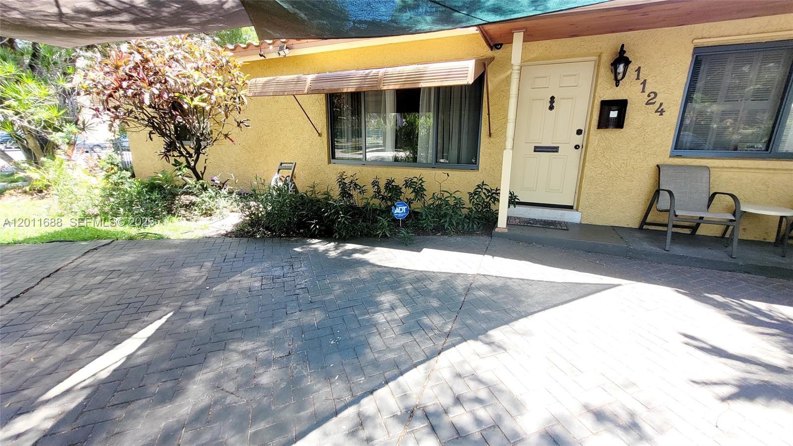 The exterior of a yellow house features a brick driveway, a front door, and landscaping — 1124 NE 7th St, Hallandale Beach FL 33009, HALLANDALE BEACH, FL