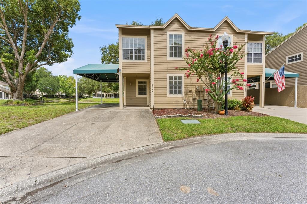 A two-story tan duplex features a green carport, a small front yard, and a tree — 9044 LAKE PLACE LANE, TAMPA, FL