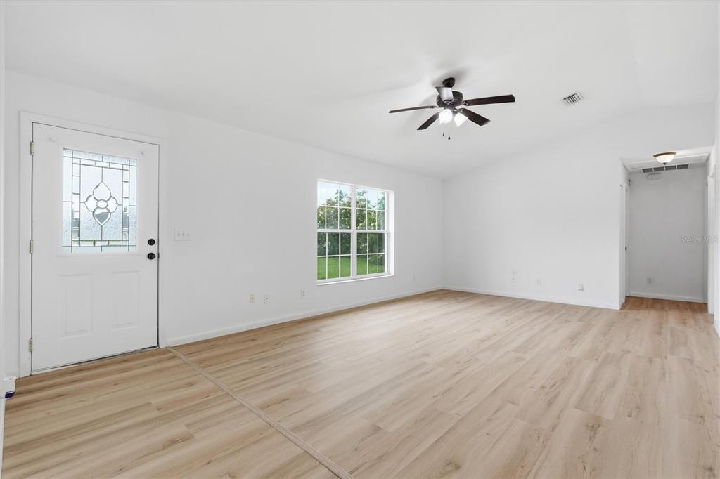 A bright living room features white walls, light wood-look flooring, a ceiling fan, and a door — 4700 SW 129TH PLACE, OCALA, FL