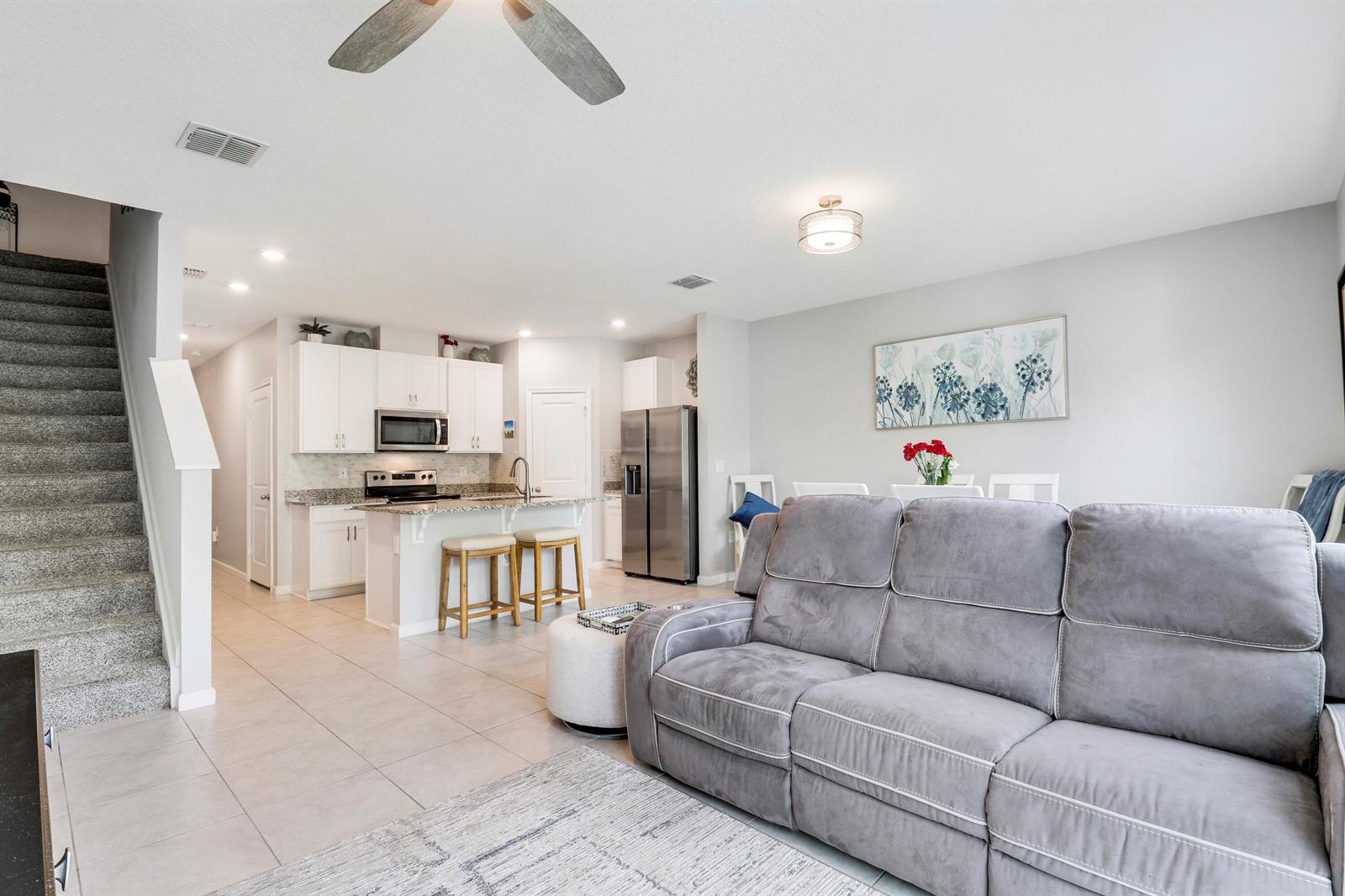 An open-concept living room and kitchen featuring a grey sofa, tiled floors, and white cabinetry — 303 SE Halifax Lane, Stuart FL 34994, STUART, FL