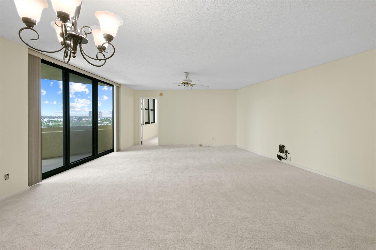 An empty living room features beige carpet, a chandelier, a ceiling fan, and sliding glass doors — 2800 N Ocean Drive # 8b, Singer Island FL 33404, SINGER ISLAND, FL