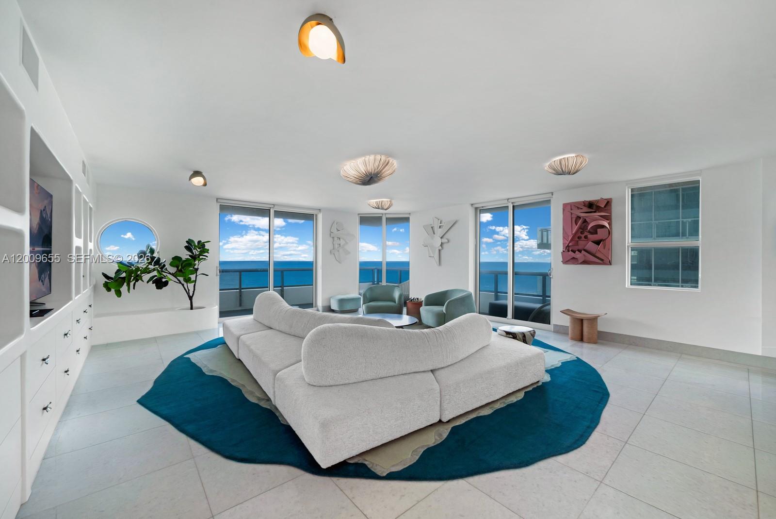A bright living room features a curved white sofa, a blue rug, and ocean views — 5801 Collins Ave # 1200, Miami Beach FL 33140, MIAMI BEACH, FL