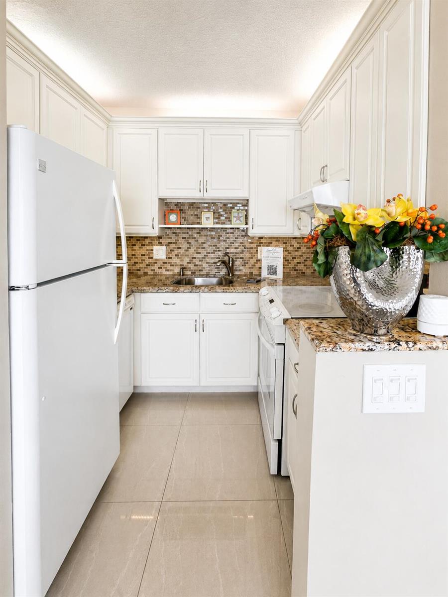 A galley kitchen features white cabinets, granite countertops, a tiled backsplash, and stainless steel appliances — 1201 S Riverside Drive # 307, Pompano Beach FL 33062, POMPANO BEACH, FL