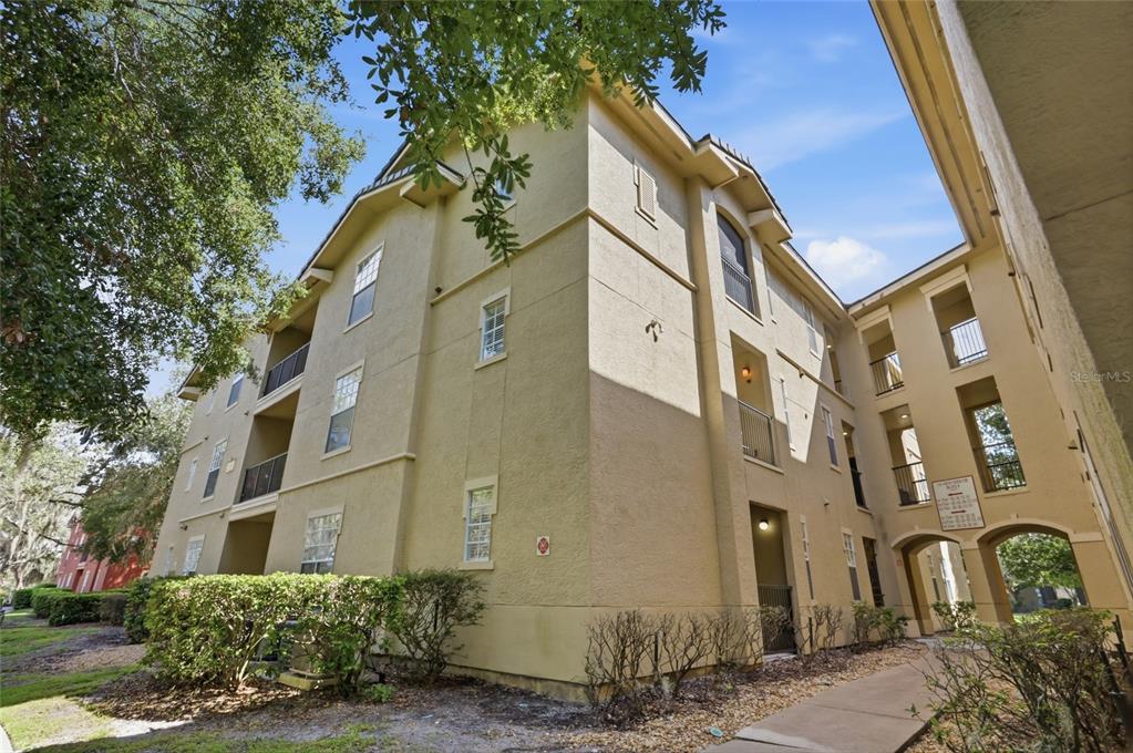 A multi-story tan apartment building features balconies, windows, and a walkway leading to an archway — 114 VISTA VERDI CIRCLE UNIT 112, LAKE MARY, FL