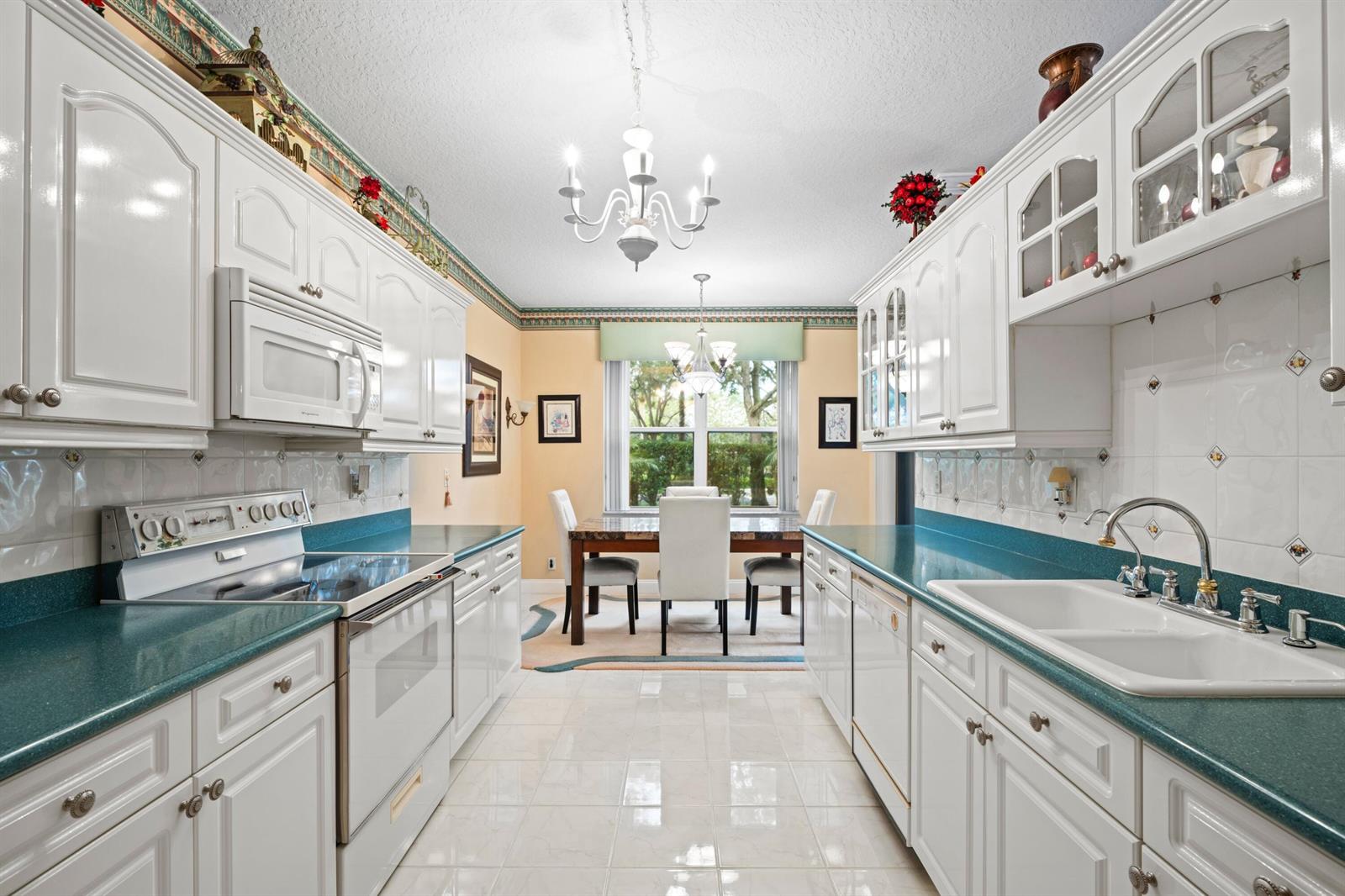 A galley kitchen with white cabinets and teal countertops leads into a dining area — 8550 Via Romana # 1, Boca Raton FL 33496, BOCA RATON, FL