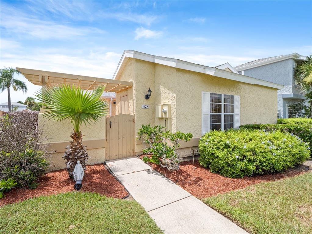 A yellow stucco house features a wooden gate, a pergola, and a concrete walkway — 1445 MALIBU CIRCLE NE UNIT 107, PALM BAY, FL