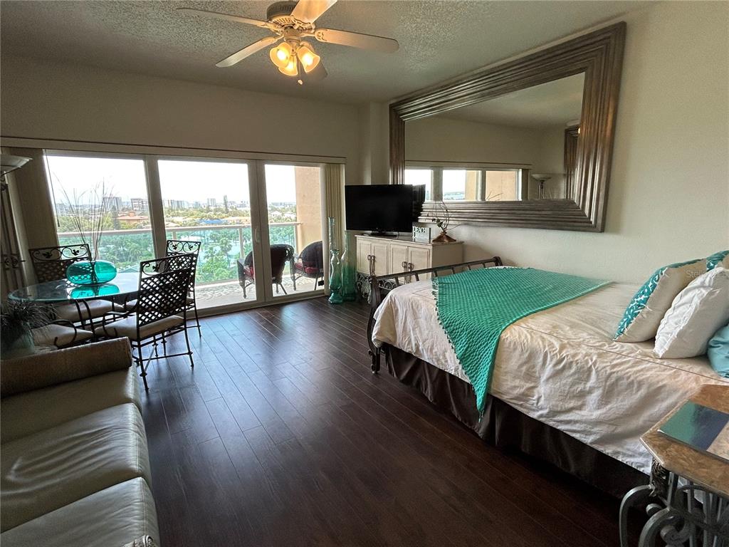 A bedroom features dark wood flooring, a bed with a teal throw, and a balcony view — 880 MANDALAY AVENUE UNIT 604, CLEARWATER BEACH, FL