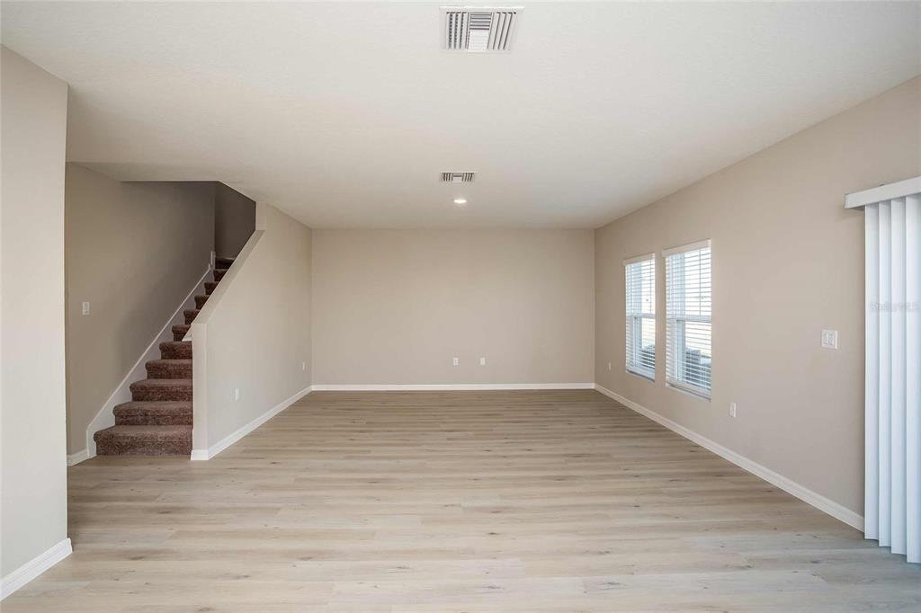 An empty living room features light wood-look flooring, beige walls, a carpeted staircase, and windows — 1807 POPPY DRIVE, LAKE HAMILTON, FL