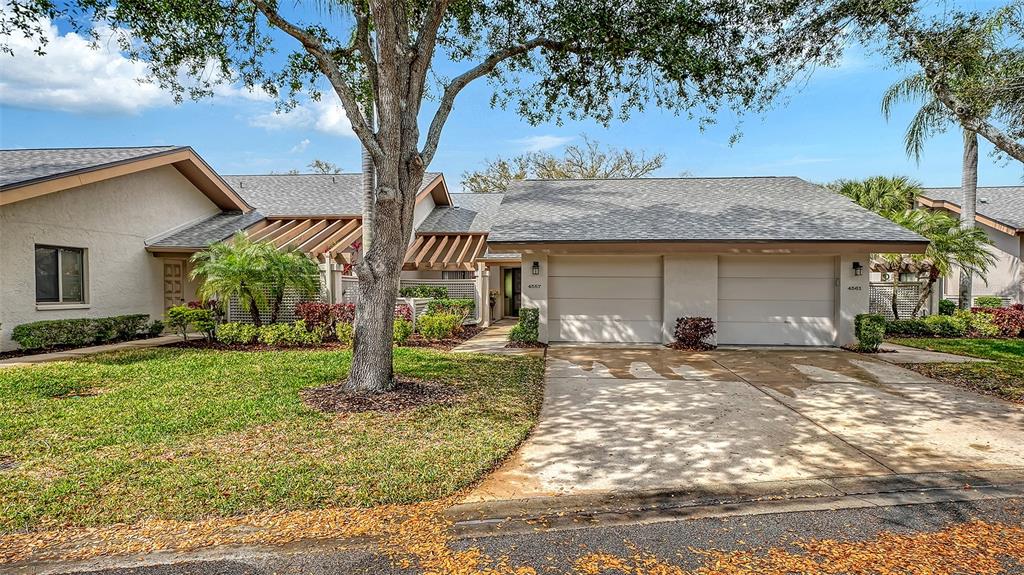 A single-story duplex with two garage doors, a large tree, and a grassy front yard — 4557 KINGSMERE UNIT 23, SARASOTA, FL