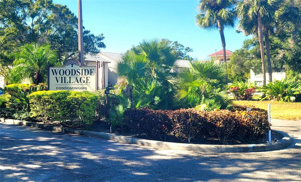 A sign for Woodside Village Condominiums stands in a landscaped area with palm trees — 4215 EAST BAY DRIVE UNIT 1508B, CLEARWATER, FL