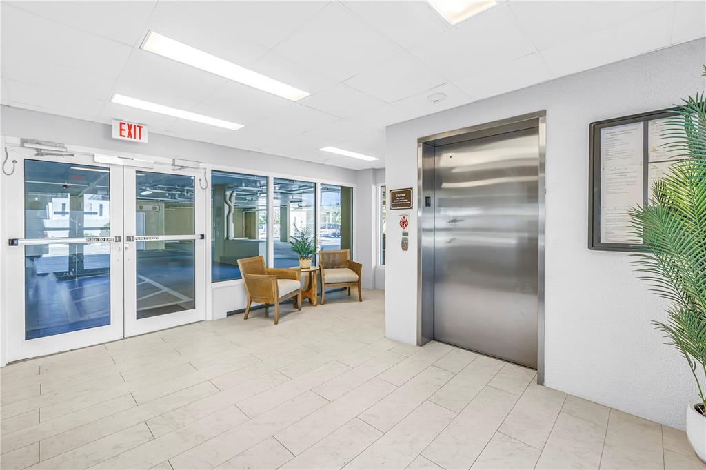 A bright lobby features a stainless steel elevator, glass exit doors, and two wicker chairs — 3805 GULF BOULEVARD UNIT 205, ST PETE BEACH, FL