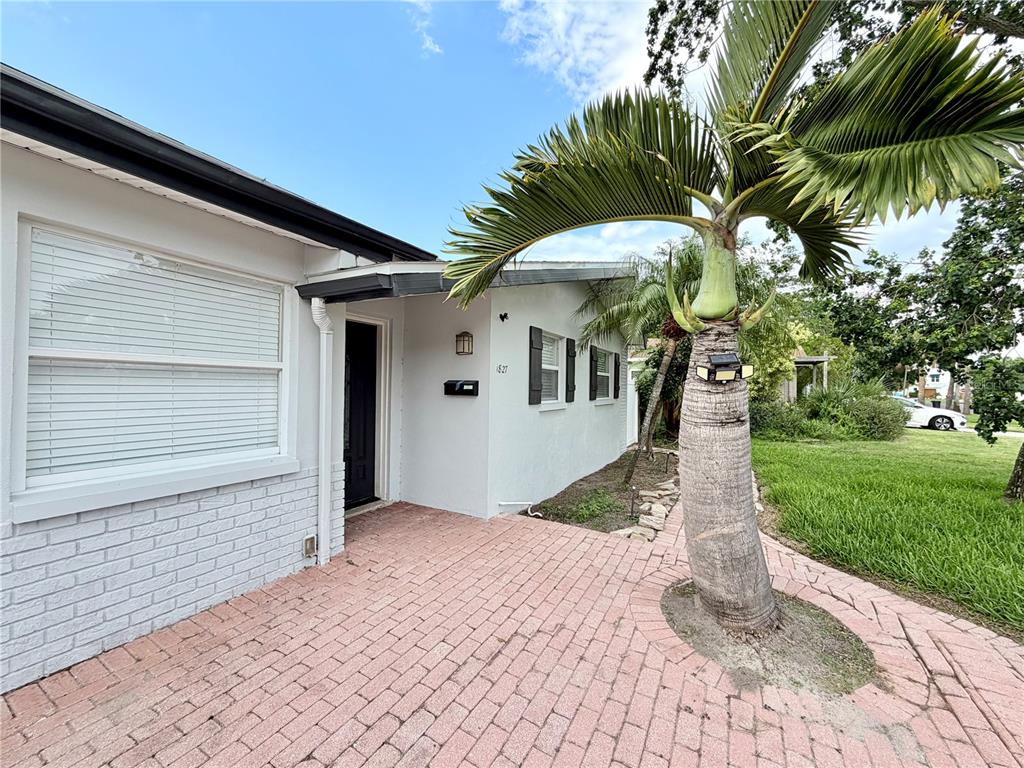 A white house exterior features a brick patio, a palm tree, and a dark door — 1827 OKLAHOMA AVENUE NE, ST PETERSBURG, FL