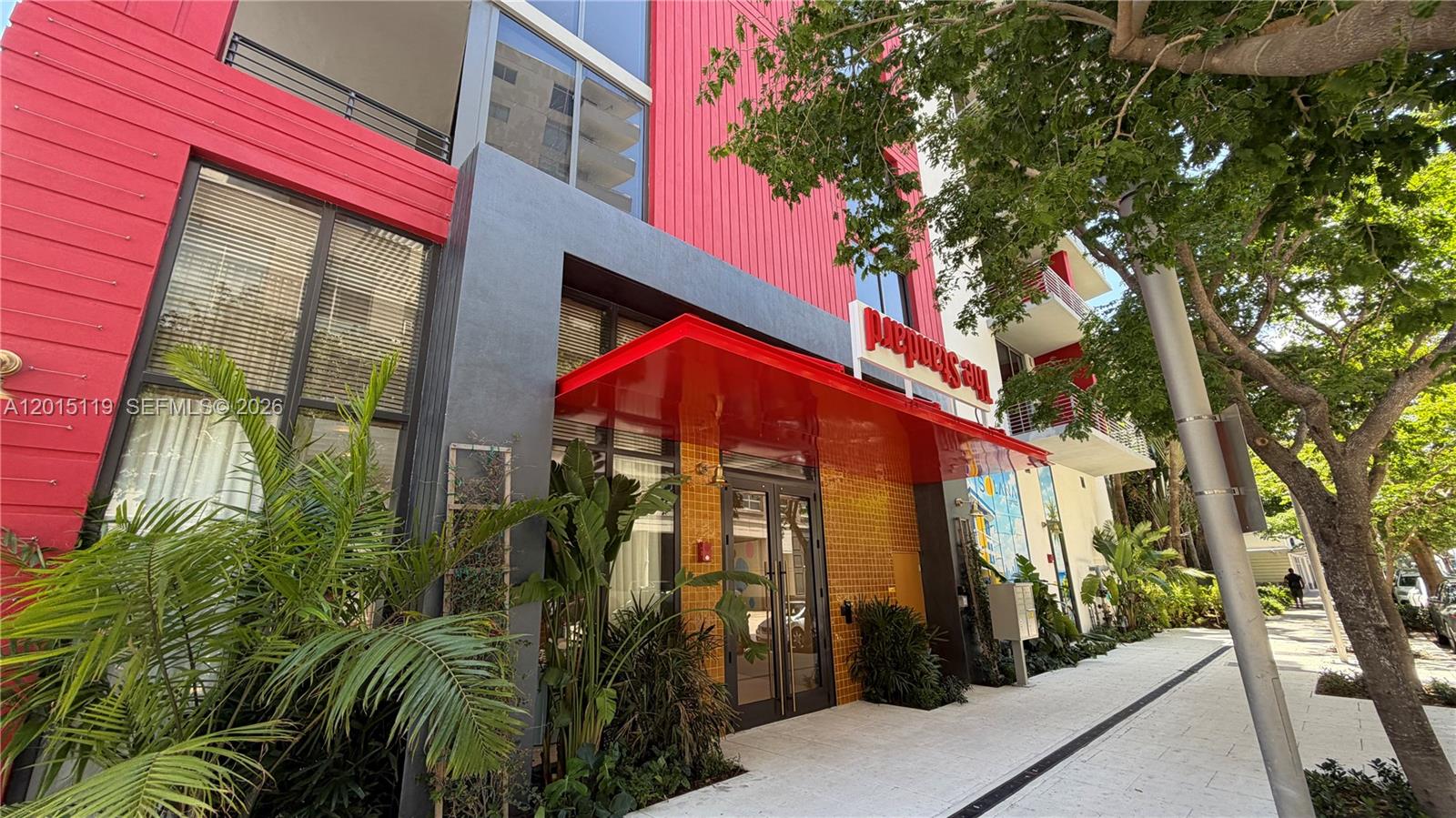 The exterior of a modern building features a red facade, glass doors, and lush landscaping — 90 NE 32nd St # 1125, Miami FL 33127, MIAMI, FL