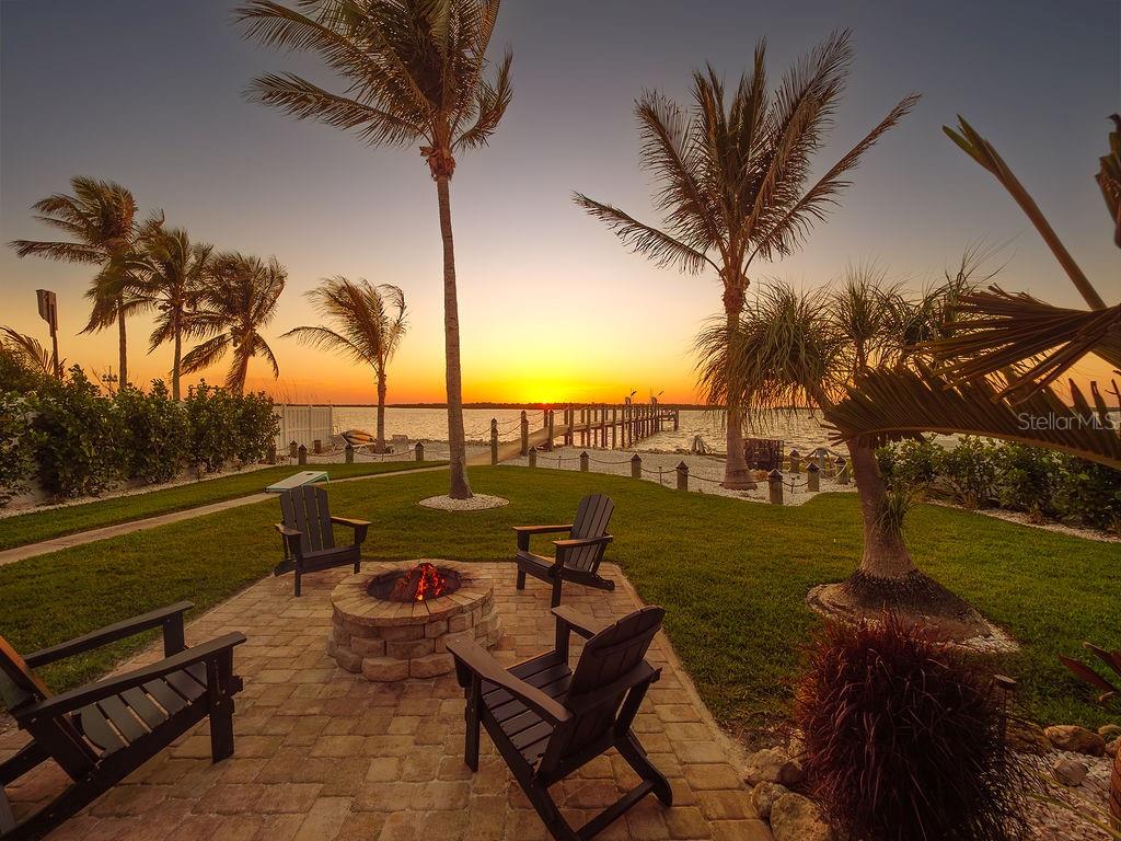 A stone fire pit with Adirondack chairs sits on a patio overlooking a sunset waterfront — 3 MICHIANA PLACE, TERRA CEIA, FL