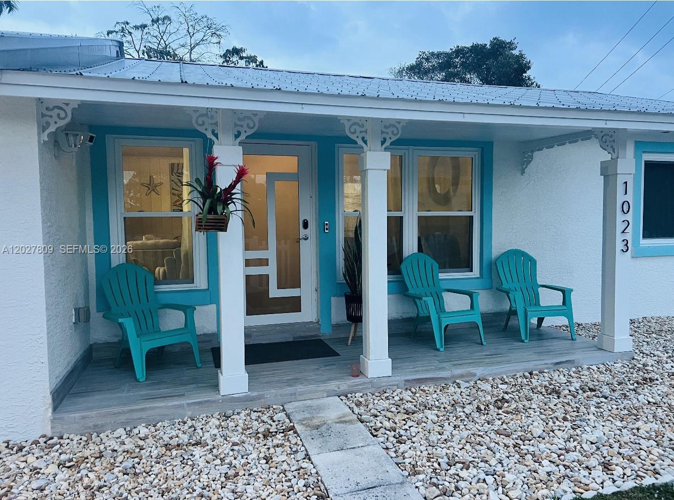 A white house exterior features a covered porch with three teal chairs and blue trim — 1023 SE 7th St, Stuart FL 34996, STUART, FL