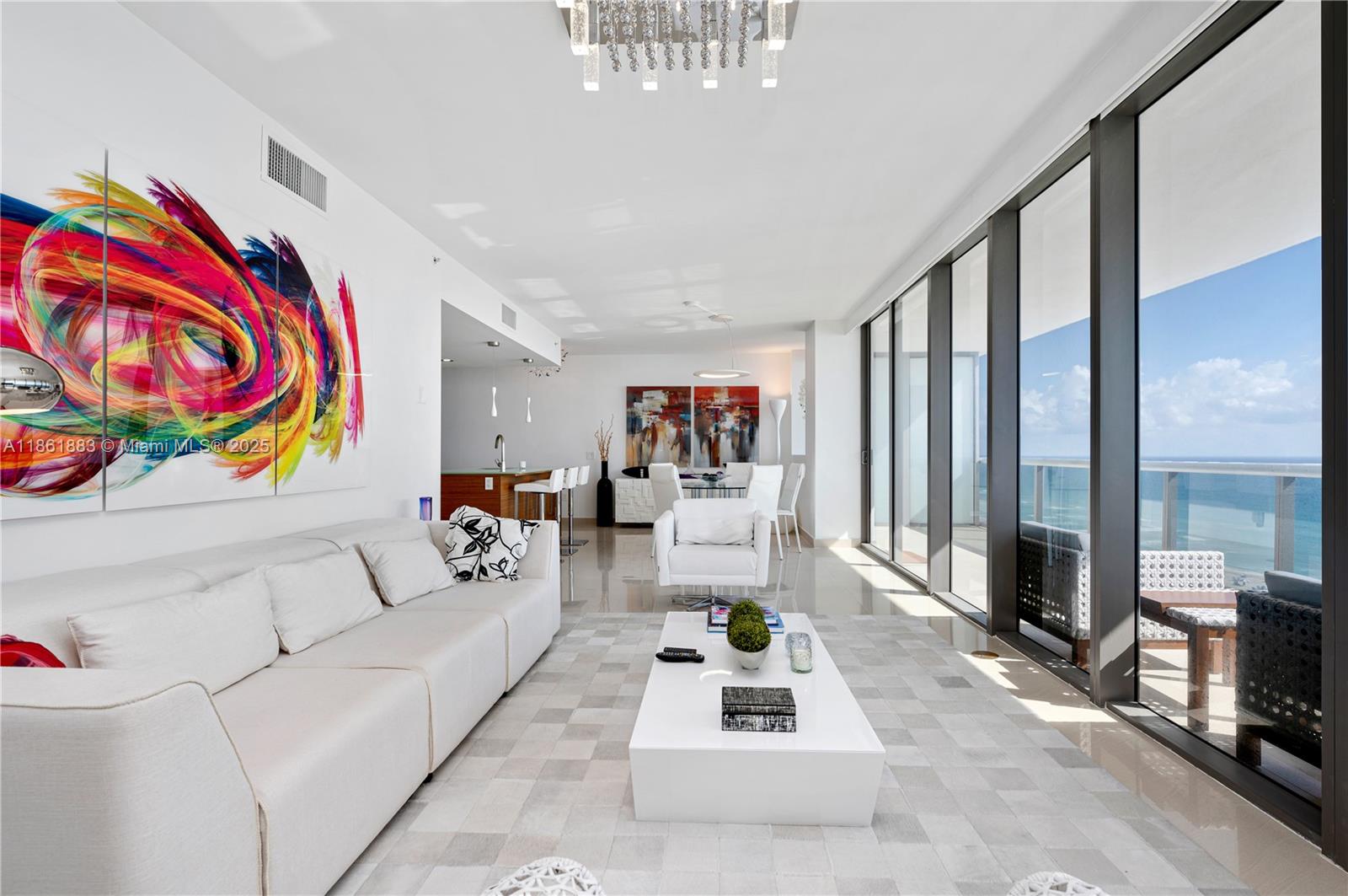 A bright living room features a white sofa, modern coffee table, and floor-to-ceiling ocean views — 5875 Collins Ave # 1202, Miami Beach FL 33140, MIAMI BEACH, FL