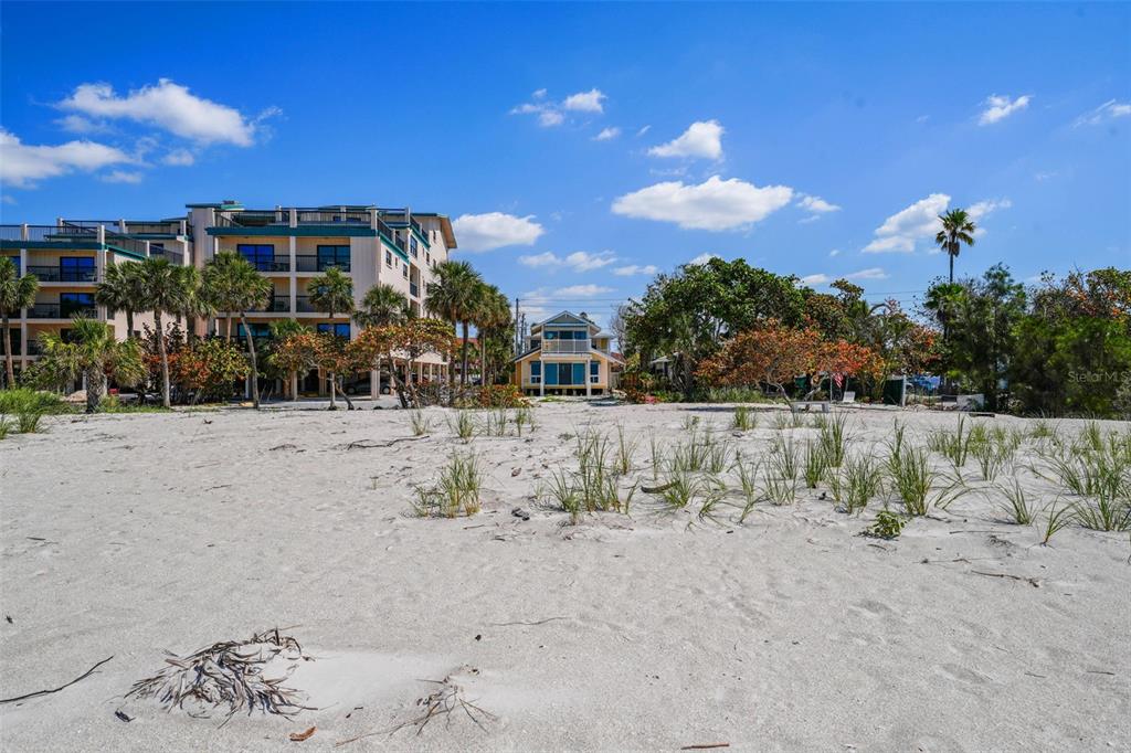 A sandy beach leads toward a multi-story apartment building and a yellow house — 13062 GULF BOULEVARD, MADEIRA BEACH, FL