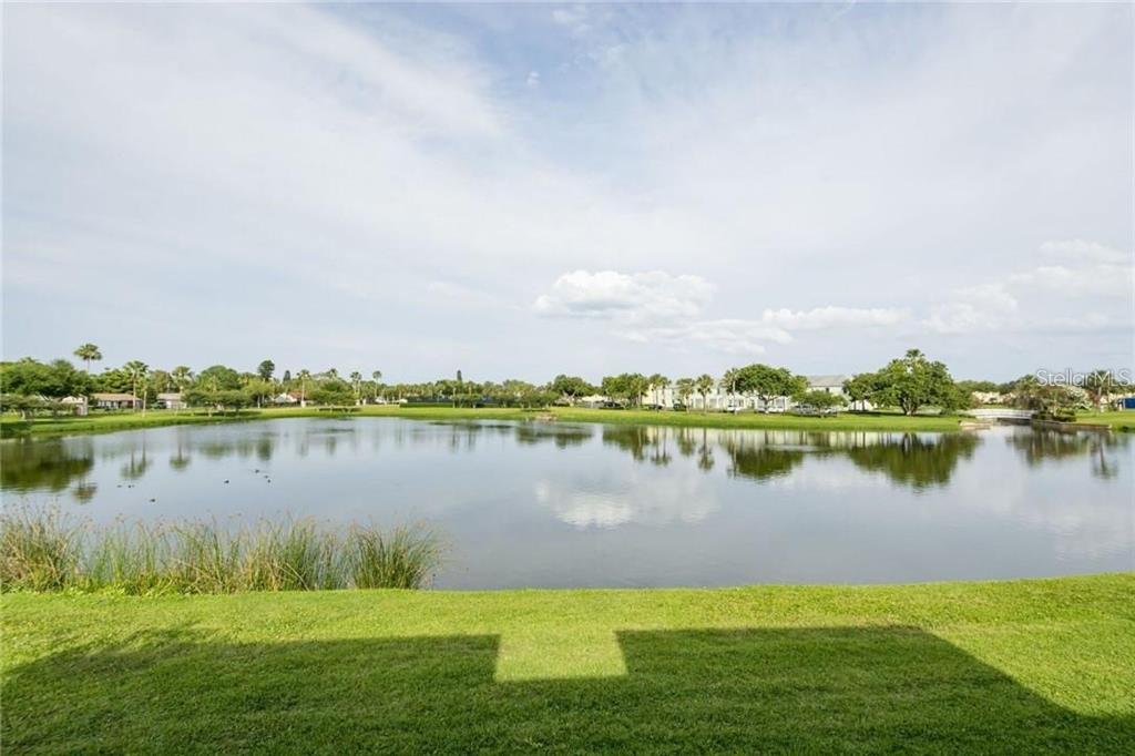 A view of a calm lake surrounded by green grass, trees, and distant houses — 4714 COBIA DRIVE SE UNIT D, ST PETERSBURG, FL