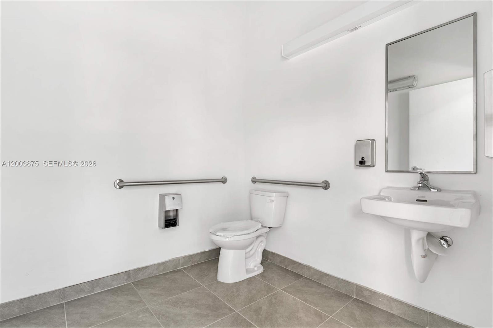 A minimalist, accessible bathroom features a white toilet, wall-mounted sink, grab bars, and tiled flooring — 9620 Stirling Rd # 101, Cooper City FL 33024, COOPER CITY, FL