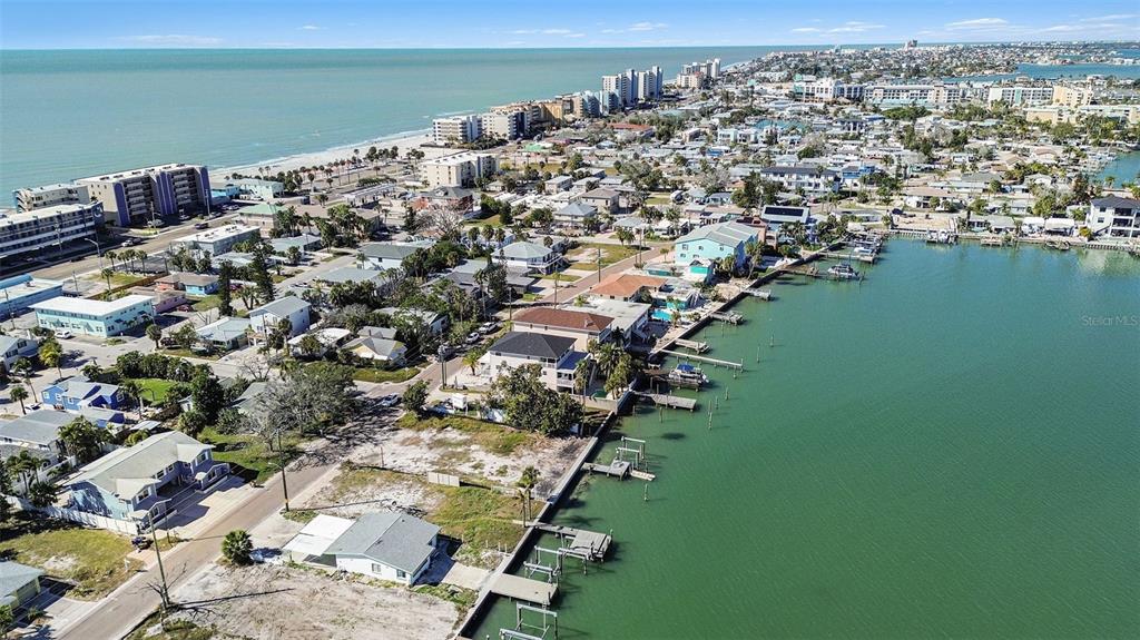 An aerial view shows a coastal neighborhood with houses, docks, and high-rise buildings near water — 14215 N BAYSHORE DRIVE, MADEIRA BEACH, FL