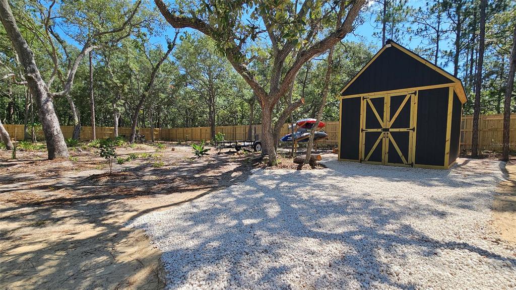 A black storage shed with yellow trim sits on a gravel pad in a wooded yard — 16266 NIGHT HERON ROAD, WEEKI WACHEE, FL
