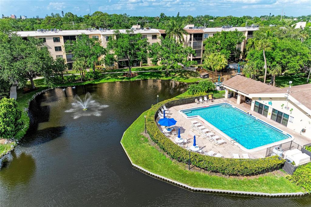 An aerial view shows a swimming pool and clubhouse beside a pond with a fountain — 625 30TH AVENUE W UNIT G203, BRADENTON, FL