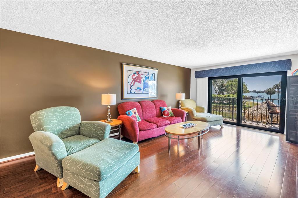 A living room with a red sofa, patterned armchair, hardwood floors, and a waterfront balcony view — 650 ISLAND WAY UNIT 204, CLEARWATER BEACH, FL