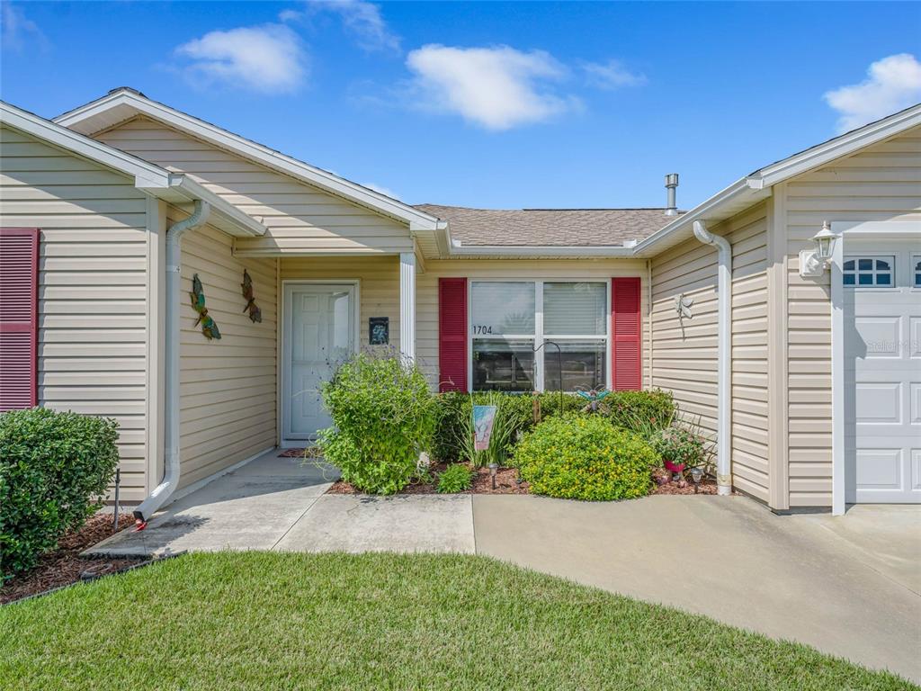 The exterior of a beige, single-story house features a front entrance, a window, and landscaping — 1704 LISETTE WAY, THE VILLAGES, FL