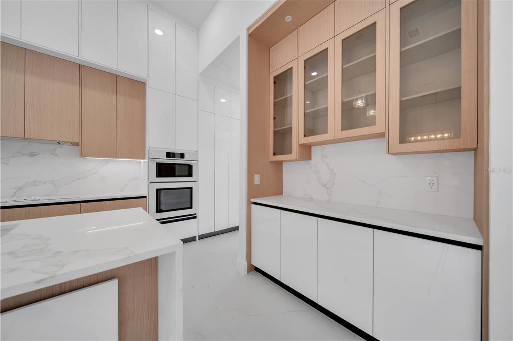 A modern kitchen features white cabinets, light wood accents, marble countertops, and built-in wall ovens — 1114 SW 8TH PL, CAPE CORAL, FL