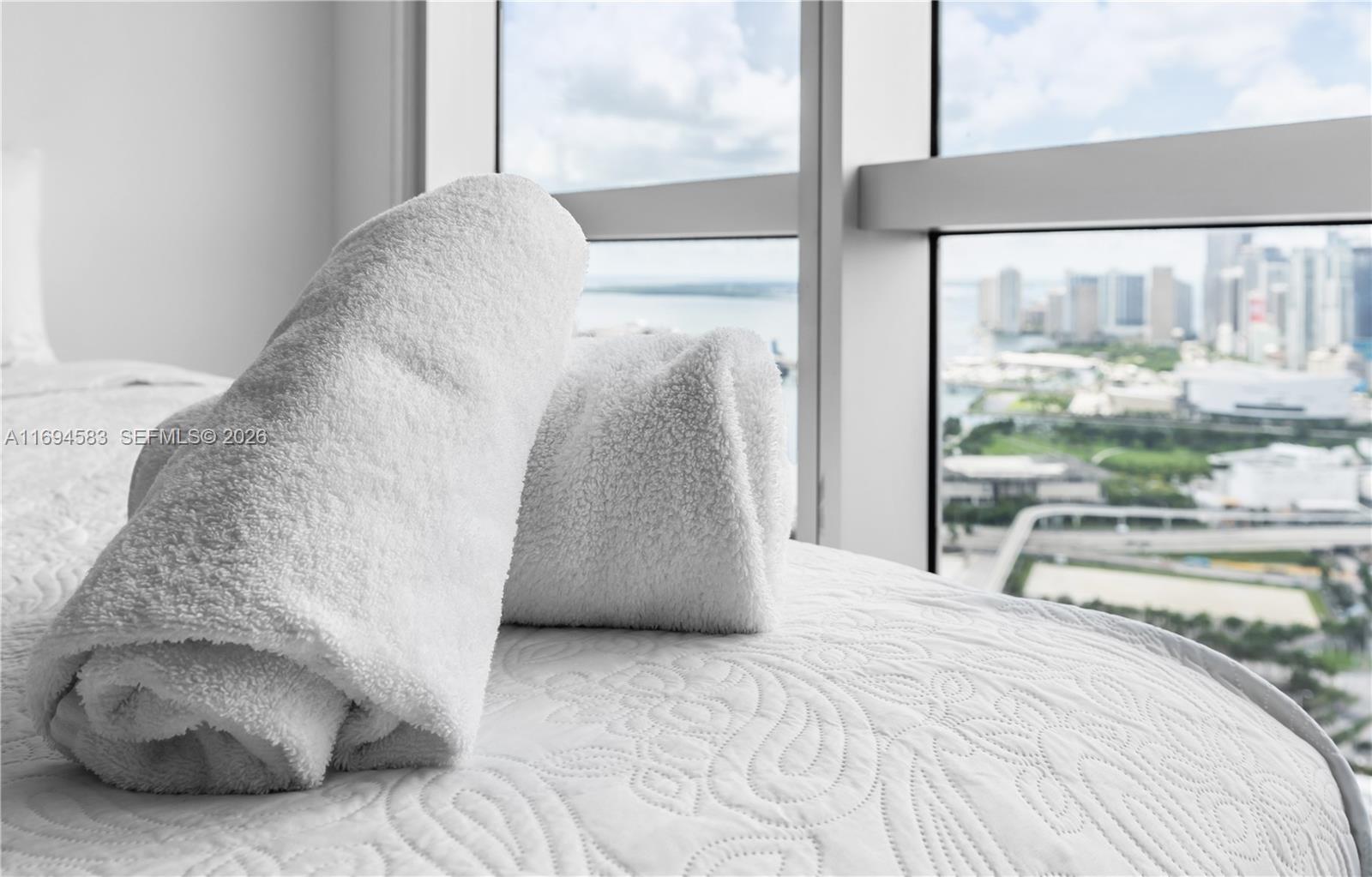 Two rolled white towels sit on a bed beside a window overlooking a city skyline — 1750 N Bayshore Dr # 5014, Miami FL 33132, MIAMI, FL