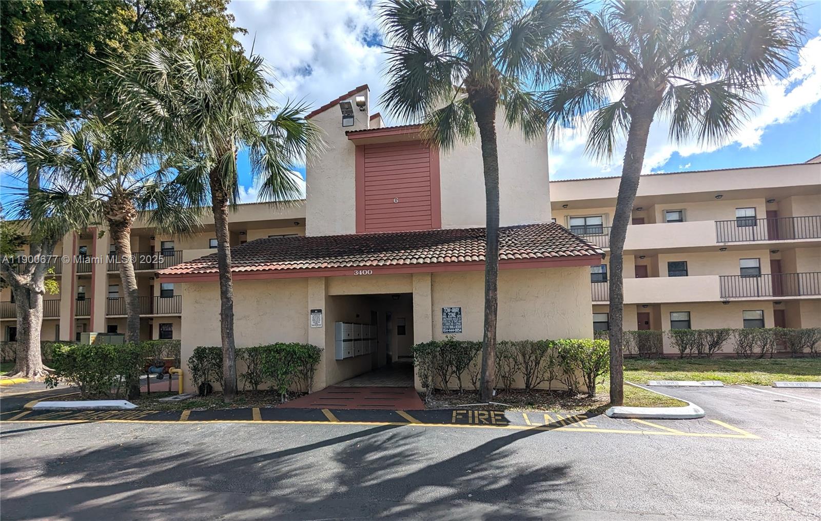 The exterior of a multi-story apartment building features a tiled roof, palm trees, and parking — 3400 Foxcroft Rd # 115, Miramar FL 33025, MIRAMAR, FL