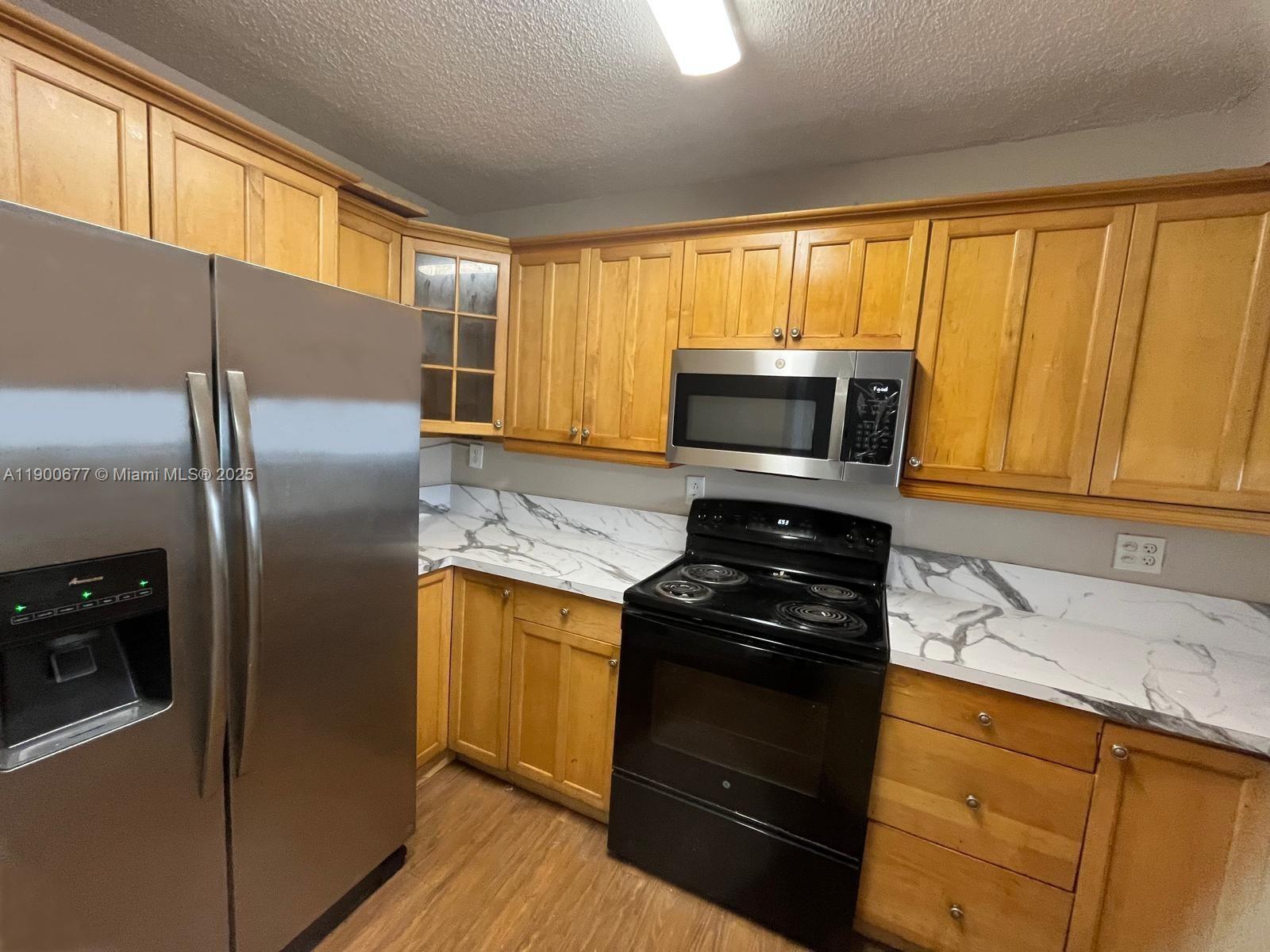 A kitchen features light wood cabinets, marble-patterned countertops, and black and stainless steel appliances — 3400 Foxcroft Rd # 115, Miramar FL 33025, MIRAMAR, FL