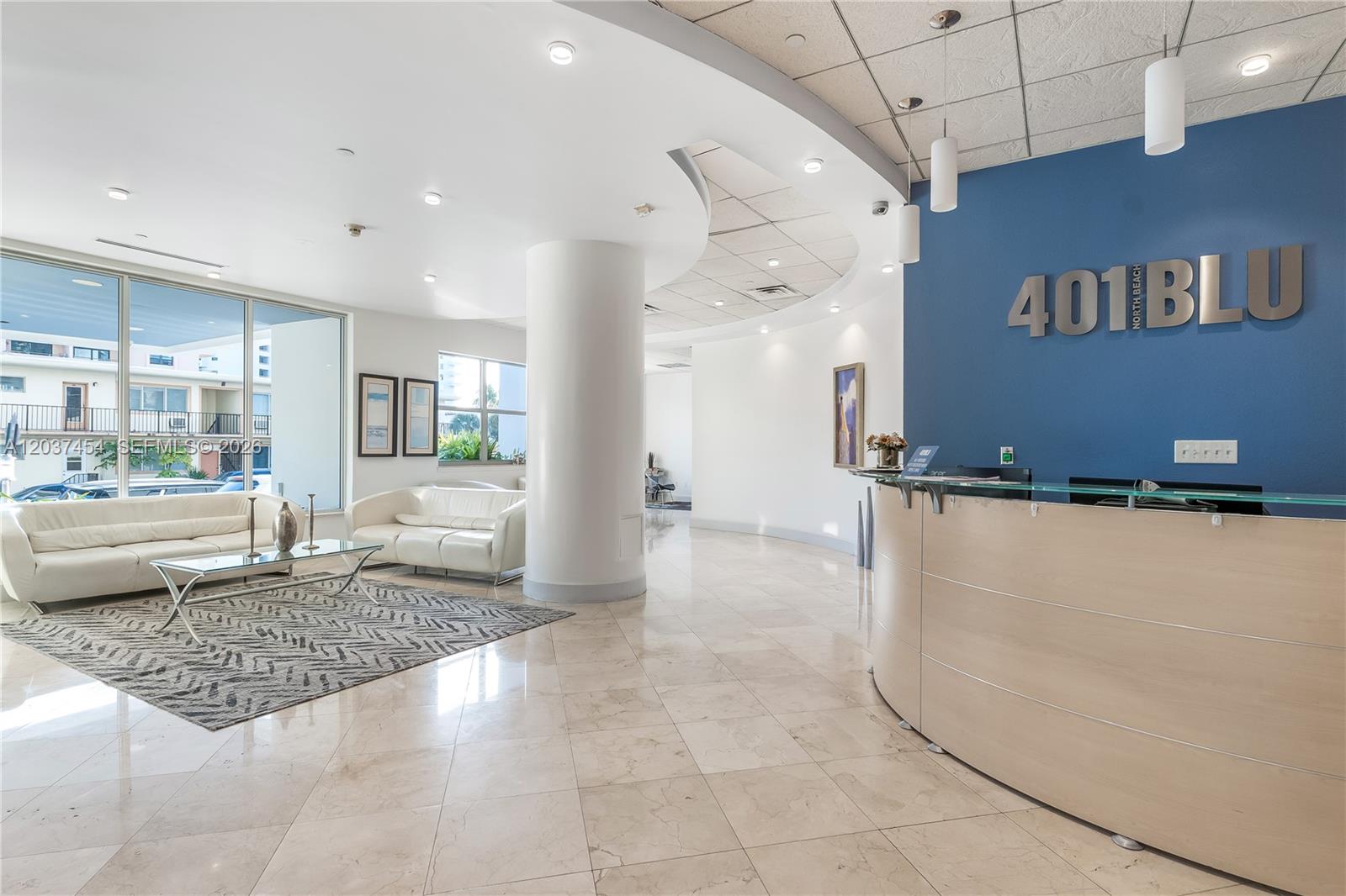 A modern lobby features a curved reception desk, white leather sofas, and a blue accent wall — 401 69th St # 313, Miami Beach FL 33141, MIAMI BEACH, FL