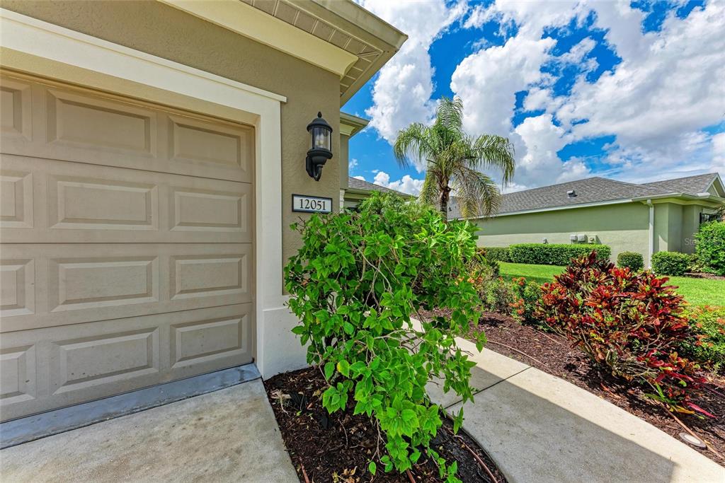 A beige garage door sits beside a green bush and a house number plaque — 12051 FOREST PARK CIRCLE, BRADENTON, FL