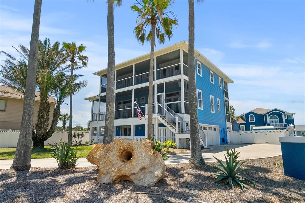 A three-story blue house with screened porches stands behind a large rock and palm trees — 1807 N CENTRAL AVENUE, FLAGLER BEACH, FL