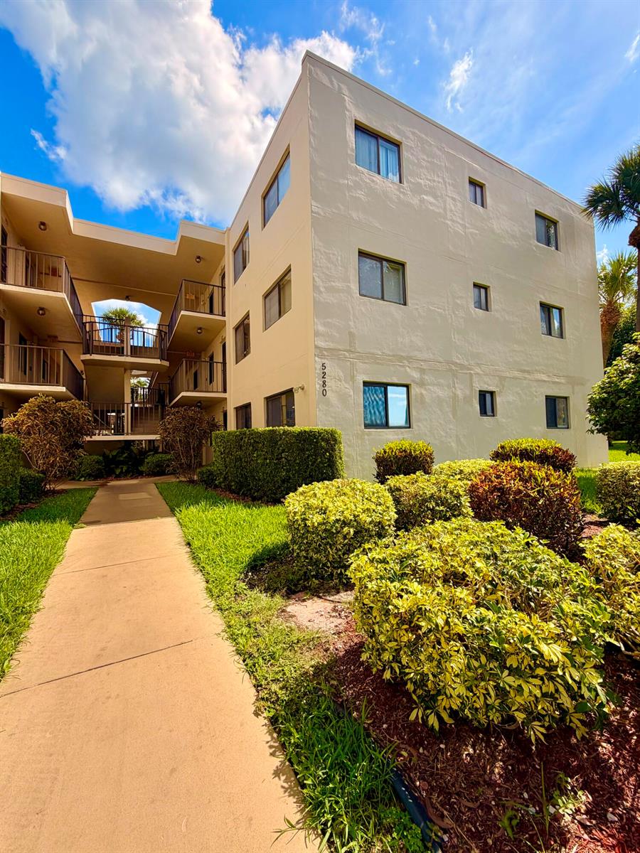 A three-story apartment building exterior with a walkway and landscaped bushes under a blue sky — 5280 Las Verdes Circle # 216, Delray Beach FL 33484, DELRAY BEACH, FL