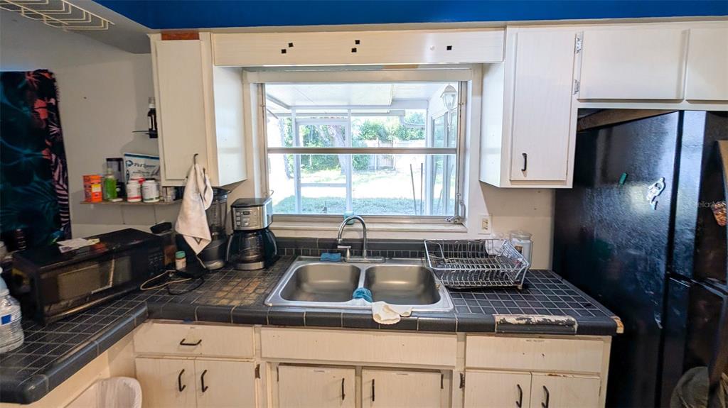A kitchen features white cabinets, a tiled countertop, a double sink, and a window view — 1590 PEACEFUL LANE N, CLEARWATER, FL