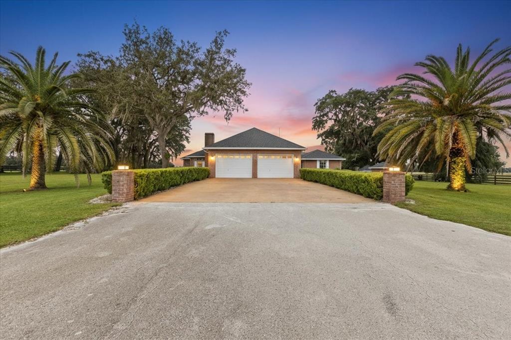 A two-car garage with a paved driveway flanked by hedges and palm trees at sunset — 10151 NW 70TH STREET, CHIEFLAND, FL