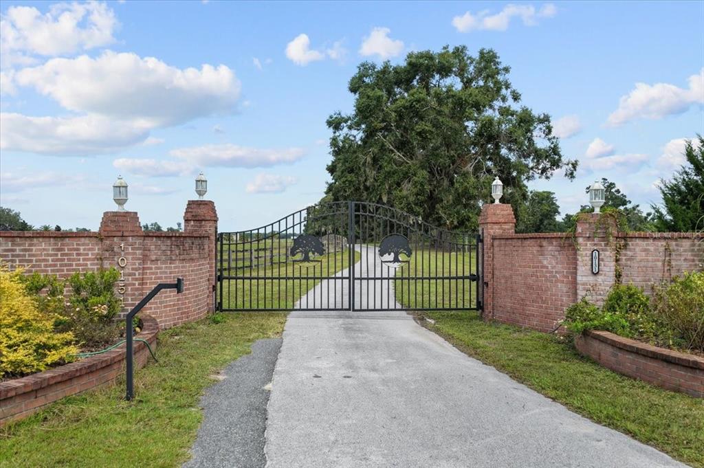 A paved driveway leads through a black metal gate flanked by brick pillars and walls — 10151 NW 70TH STREET, CHIEFLAND, FL
