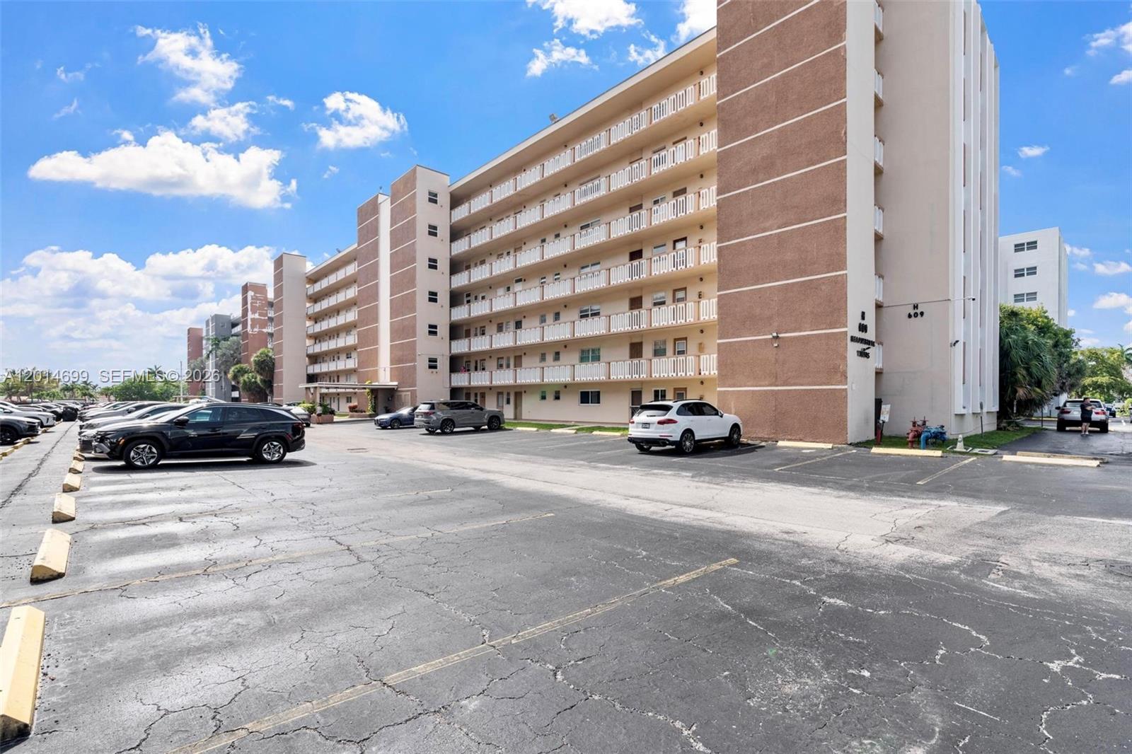 A multi-story apartment building with a parking lot in the foreground under a blue sky — 609 NE 14th Ave # 405, Hallandale Beach FL 33009, HALLANDALE BEACH, FL