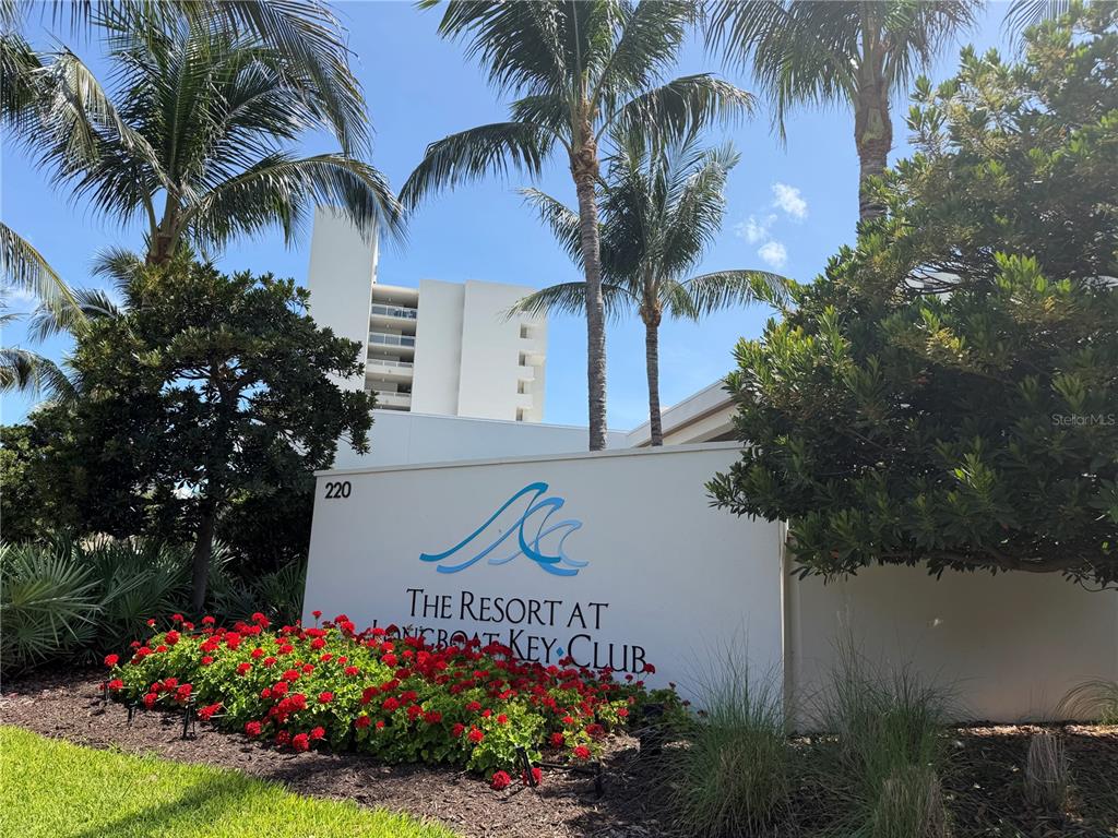 A white sign with blue wave graphics stands before a tall building and palm trees — 210 SANDS POINT ROAD UNIT 2402, LONGBOAT KEY, FL