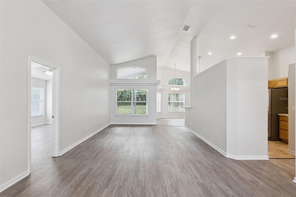 An empty living room with vaulted ceilings, gray wood-look flooring, and an adjacent kitchen area — 5483 GLEN OAK PLACE, SANFORD, FL