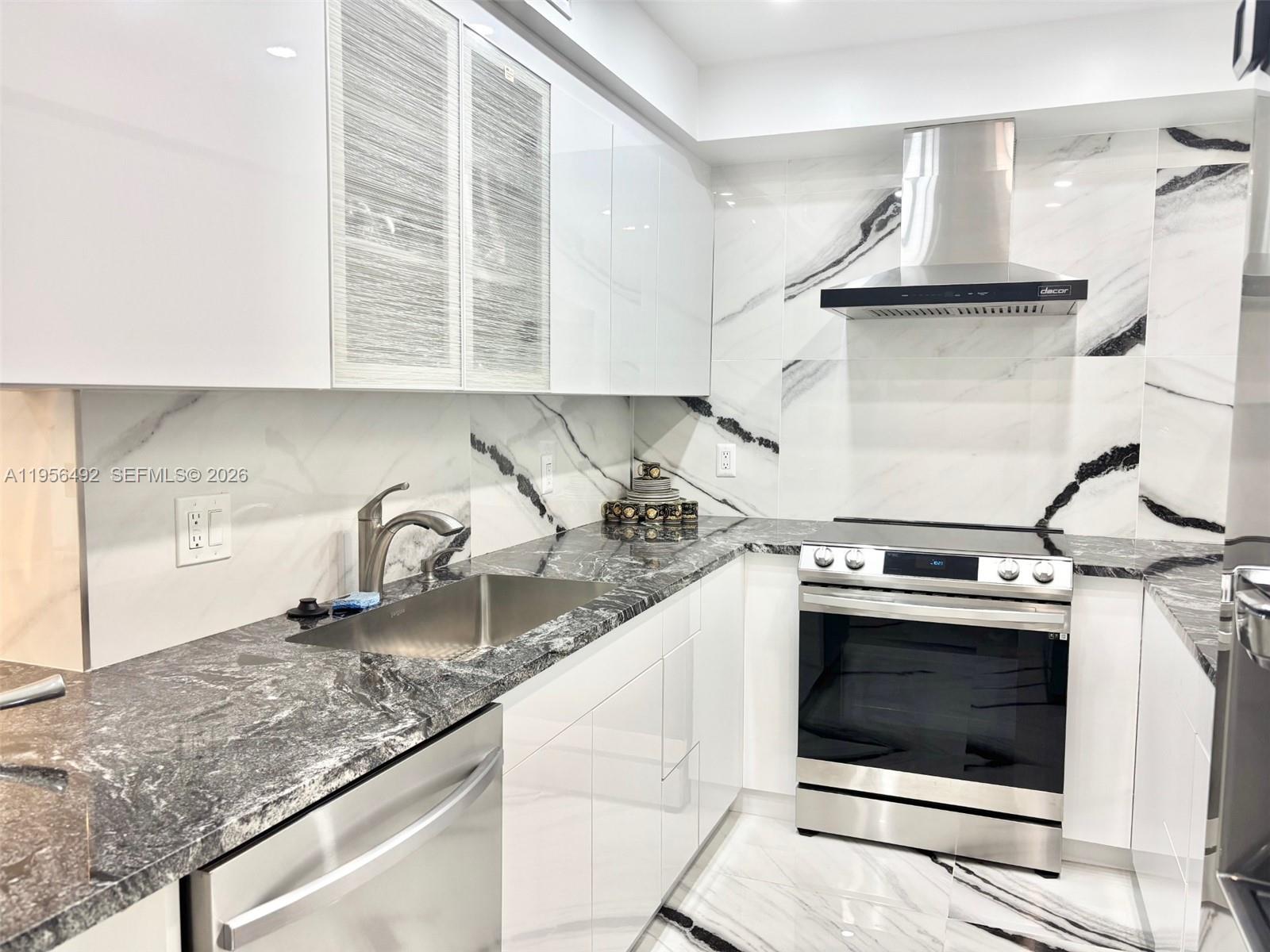 A modern kitchen features white cabinets, marble-patterned walls, and dark granite countertops — 1965 S Ocean Dr # 15B, Hallandale Beach FL 33009, HALLANDALE BEACH, FL