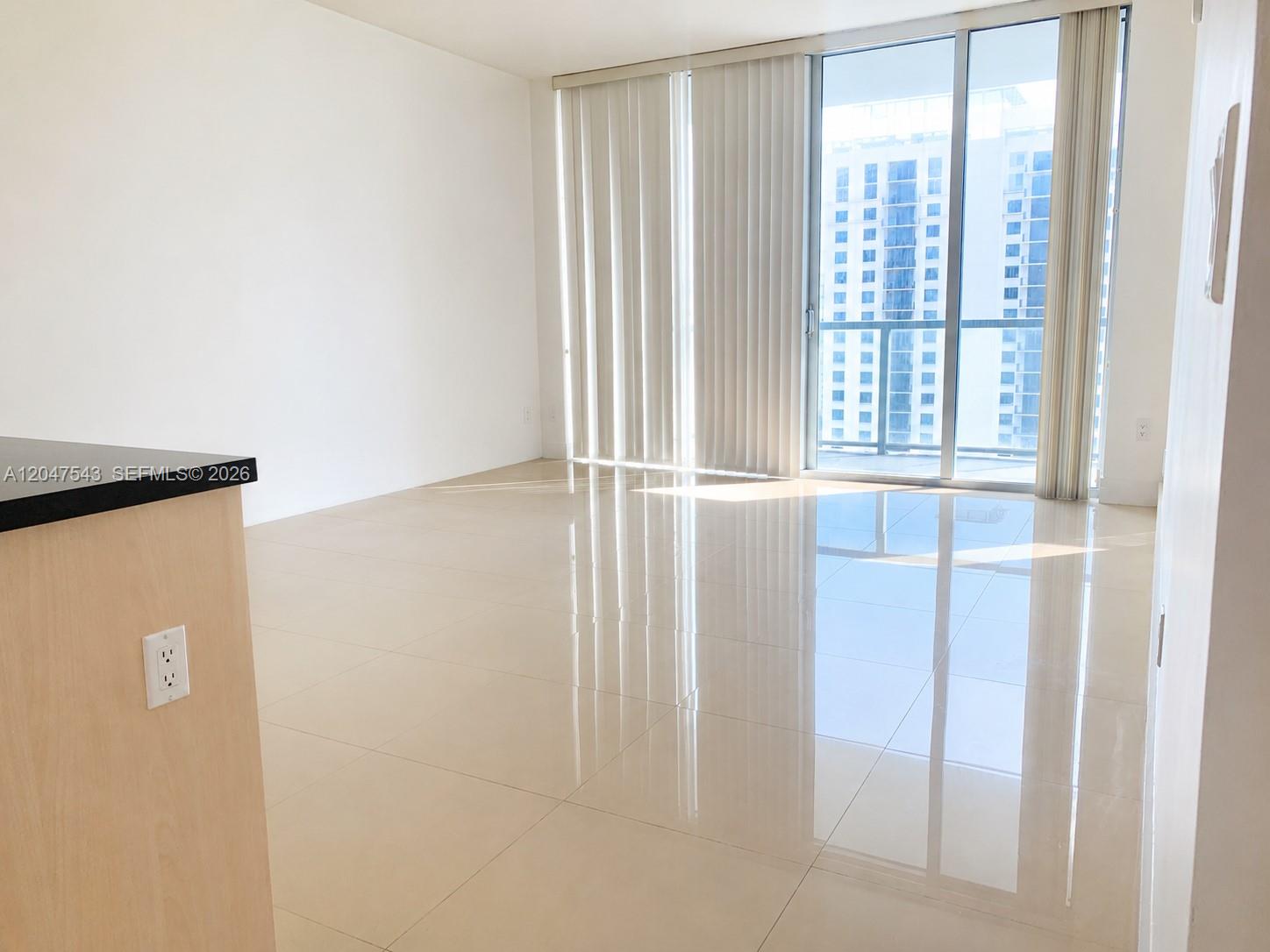An empty room features light-colored tile flooring, white walls, and floor-to-ceiling windows with vertical blinds — MIAMI, FL