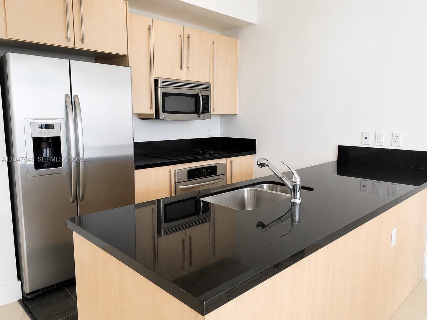 A kitchen features light wood cabinets, black countertops, stainless steel appliances, and a central island — MIAMI, FL