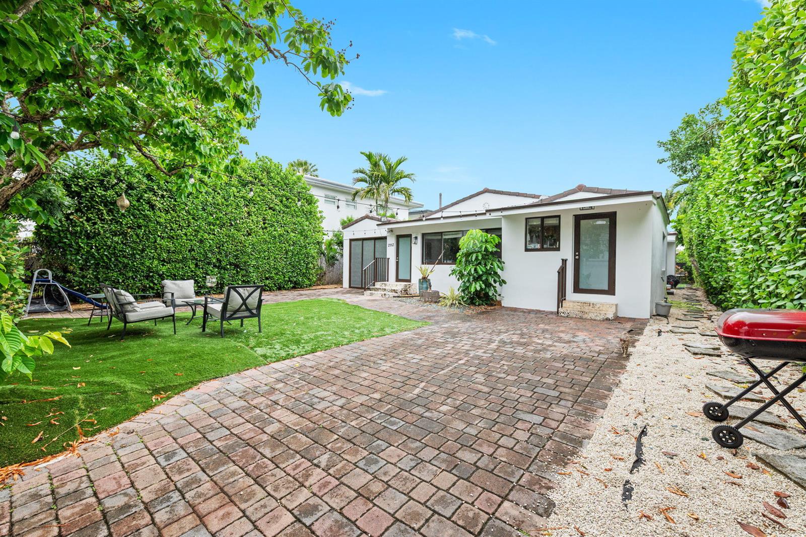 A white house exterior features a brick patio, artificial turf lawn, and lush privacy hedges — 2183 Bay Drive, Miami Beach FL 33141, MIAMI BEACH, FL