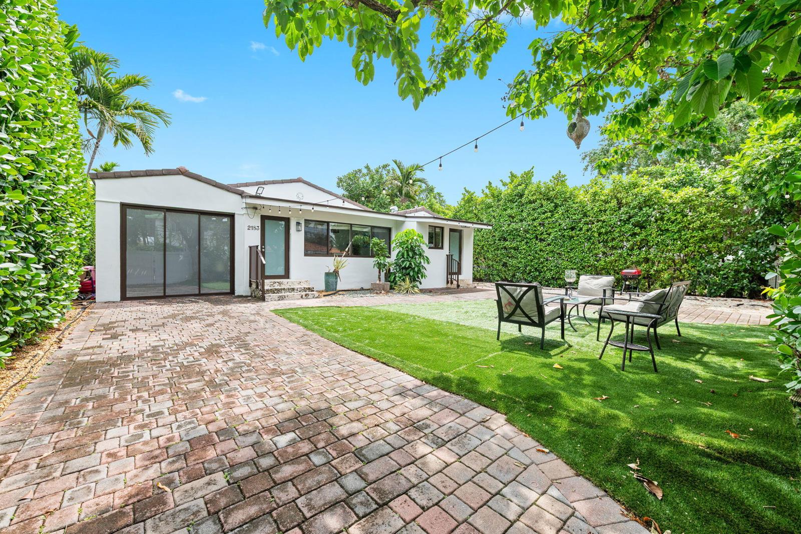 A white house with a brick patio and artificial turf lawn surrounded by tall hedges — 2183 Bay Drive, Miami Beach FL 33141, MIAMI BEACH, FL